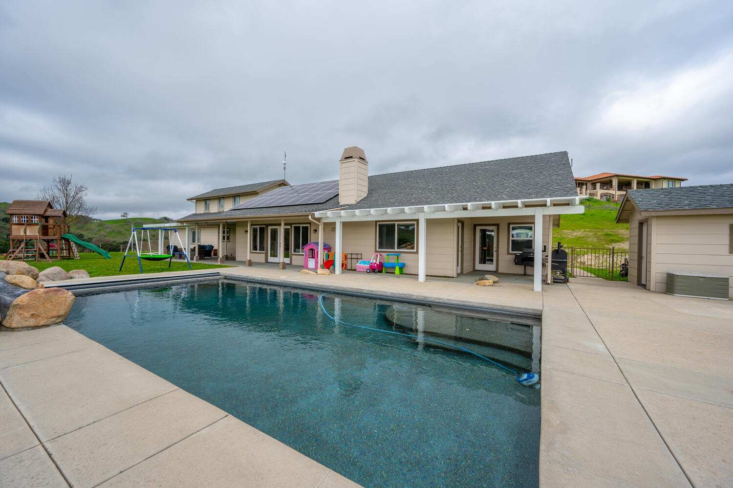Backyard of a Templeton home with a swimming pool, covered patio, rooftop solar panels and a lawn with play equipment
